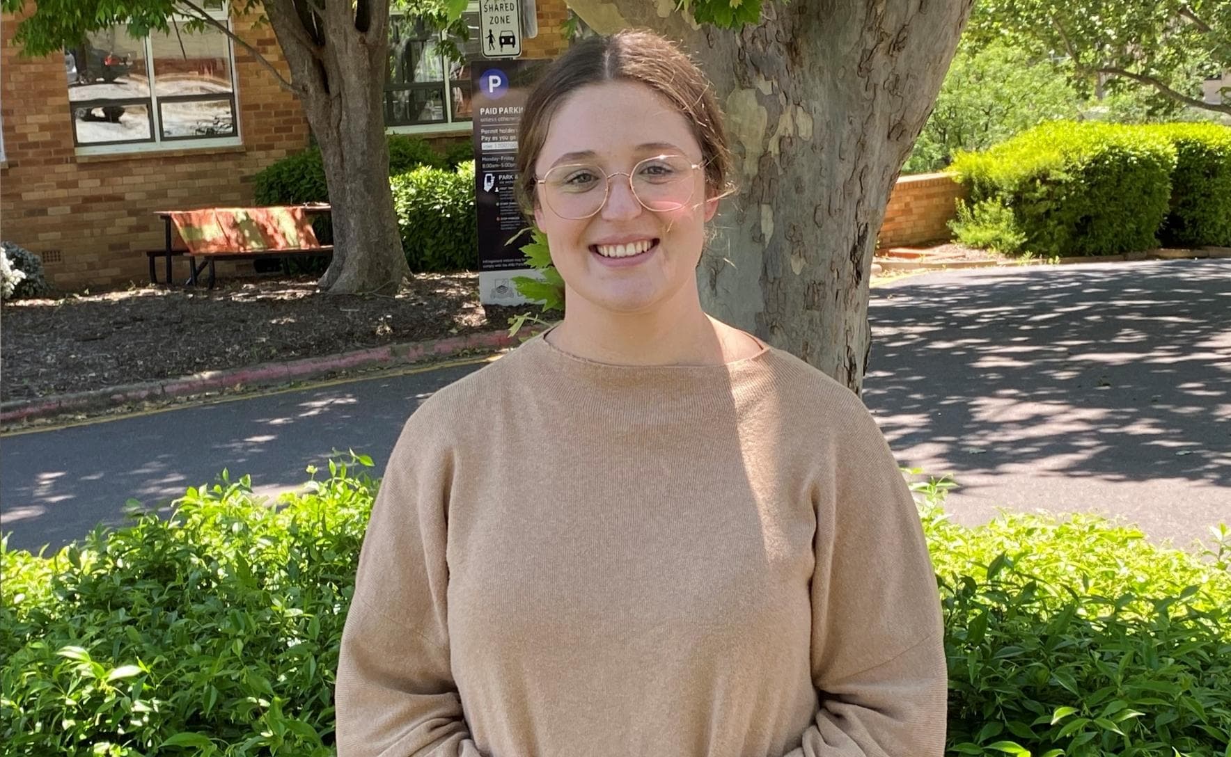 Mikayla is wearig a brown jumper and standing in front of a tree. She is smiling at the camera