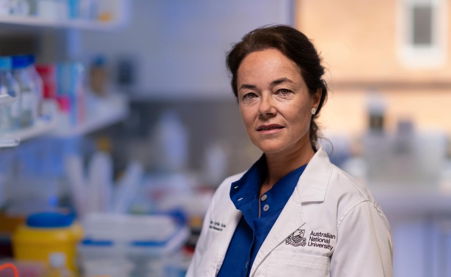 Carola is wearing a whit elab coat and blue shirt. She is looking at the camera and a blurred background of a laboratory is behind her. 