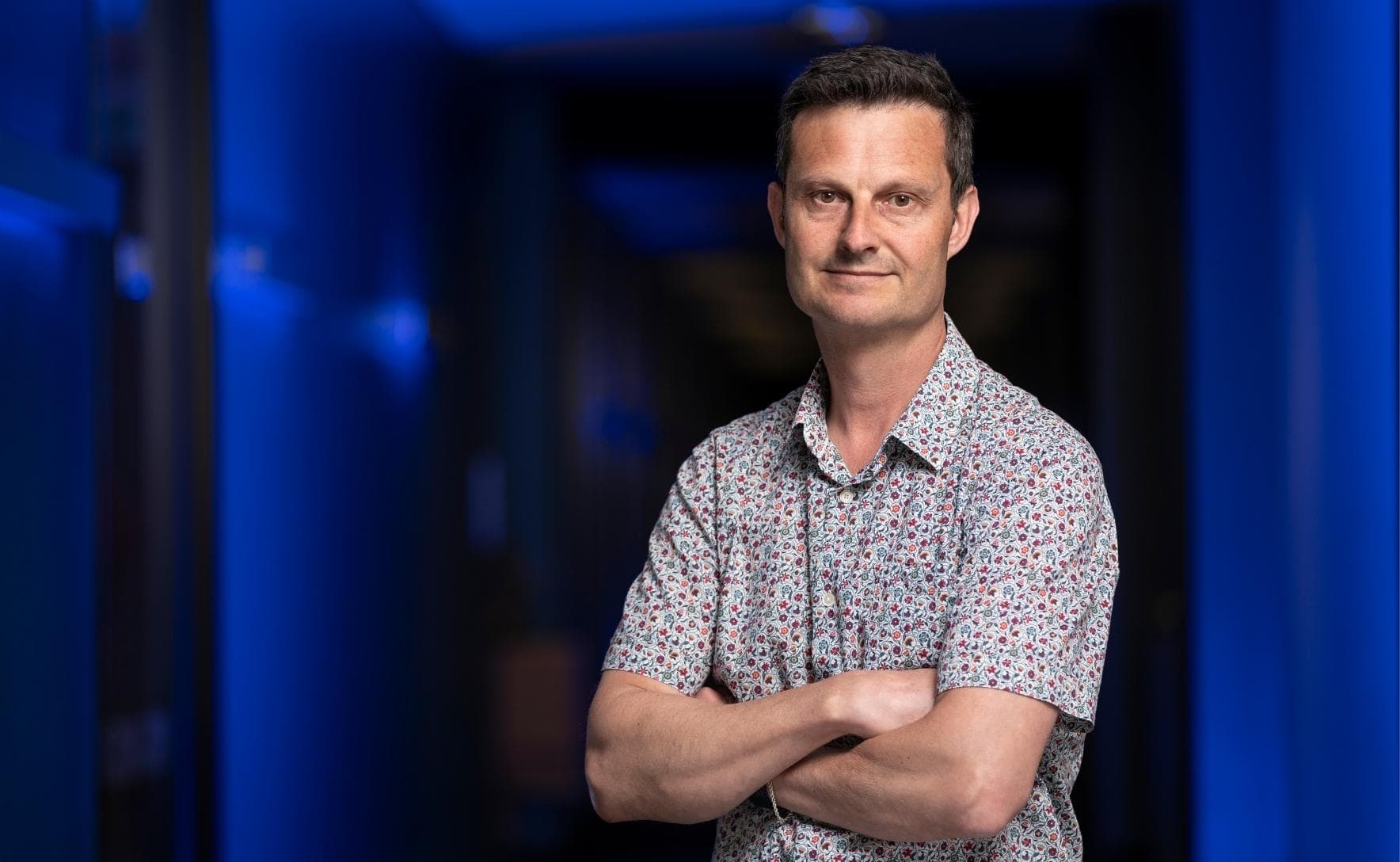 David is standing in front of a dark corridor lit by blue lights. He is wearing a floral collared shirt, with his arms crossed and smiling at the camera