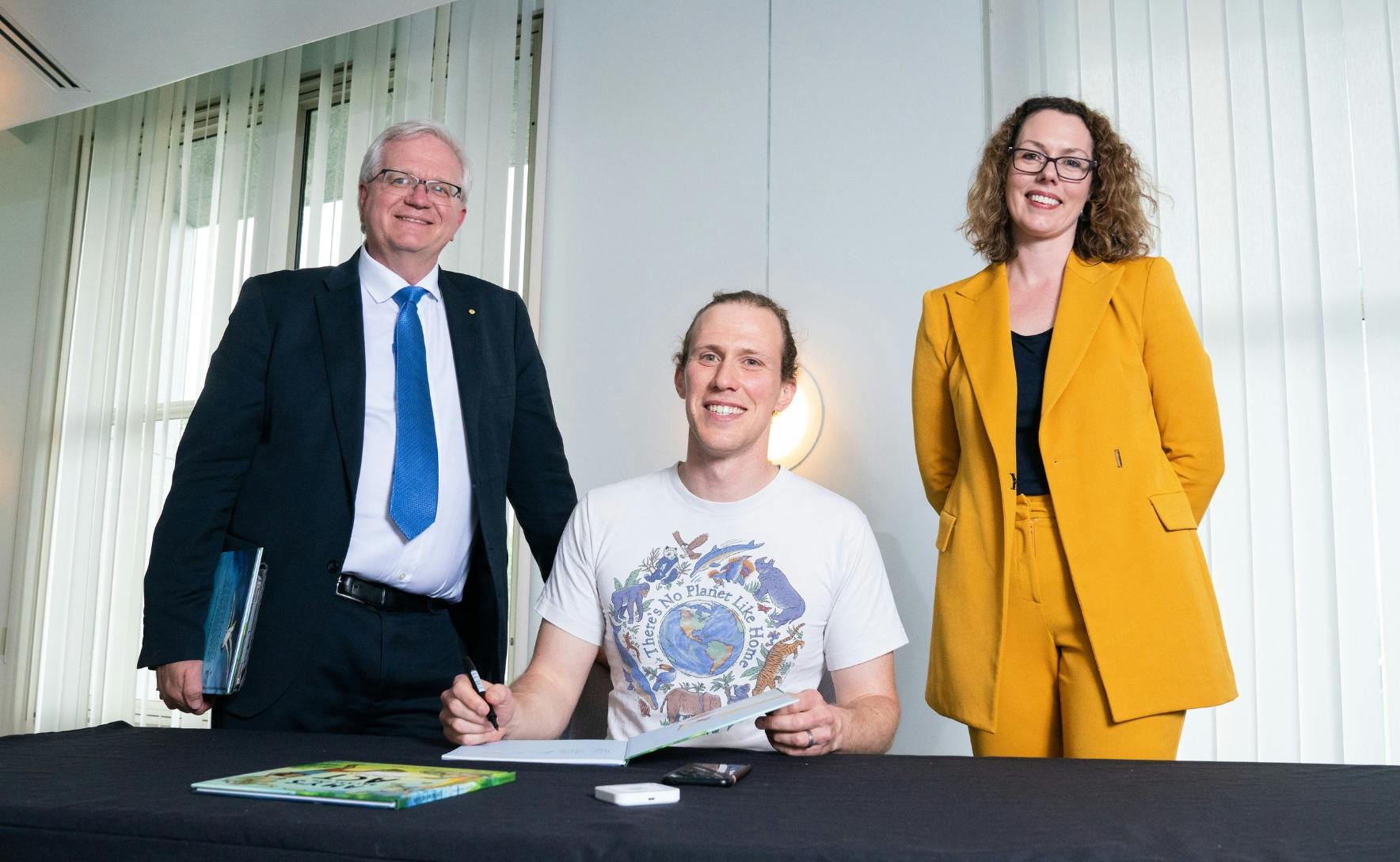 A man and a woman are standing and smiling at the camera. In between them, another man is sitting at a table and smiling while signing a book