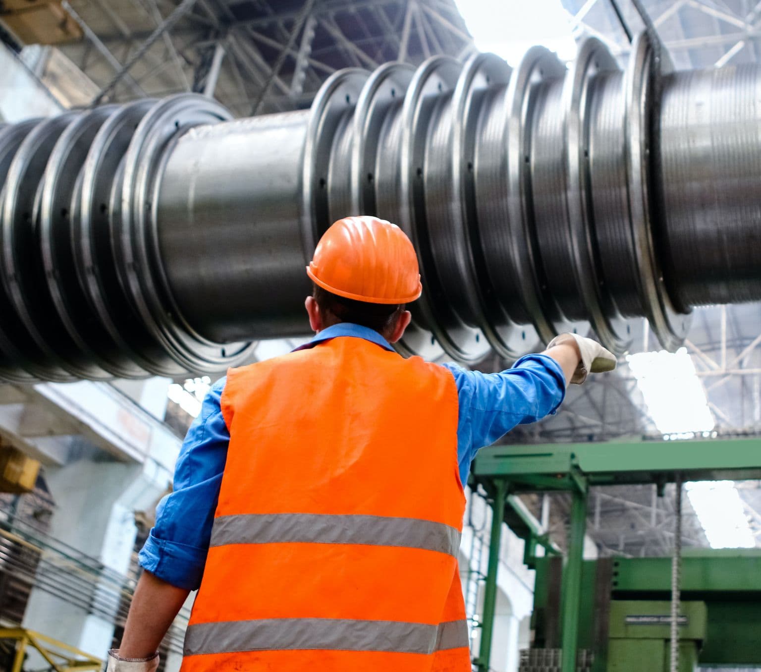 back of a man wearing an orange vest and orange safety helmet. He is reaching up to a large steel pipe in a factory