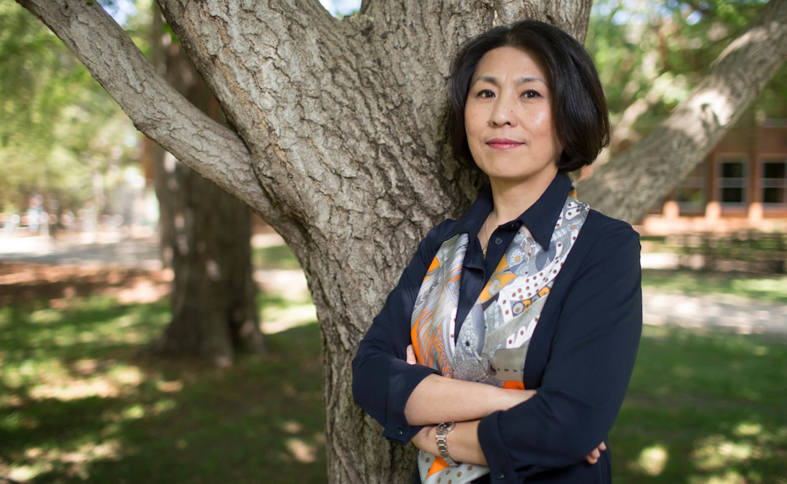 Xuemei Bai stands in front of a tree. She wears a dark blue cllard shirt and a patterned scarf.