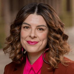 Jarrah Aguera smiles at the camera, she has shoulder length brown curly hair and is wearing a pink shirt and red jacket.