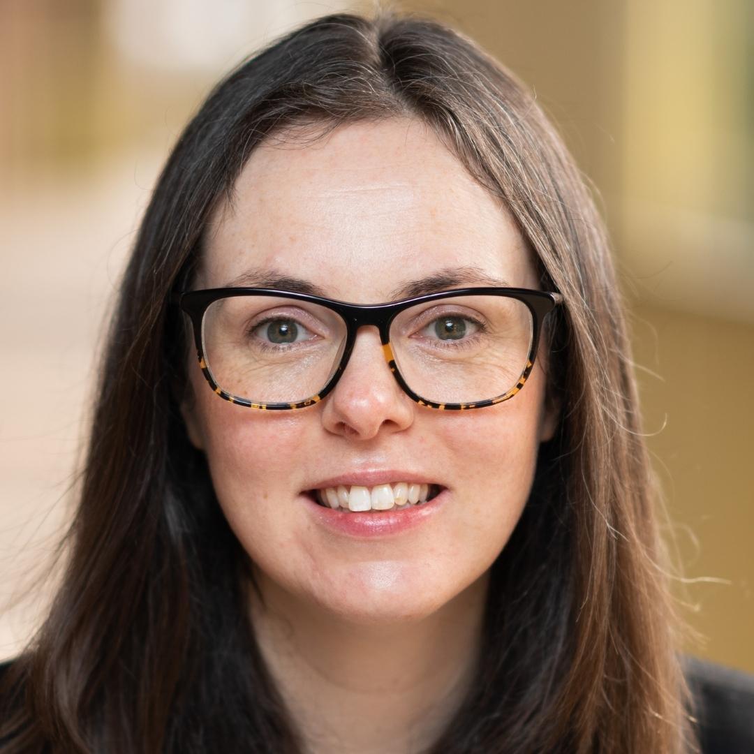 Megan Dingwall smiles at the camera, she has long brown hair and wears black and yellow glasses.