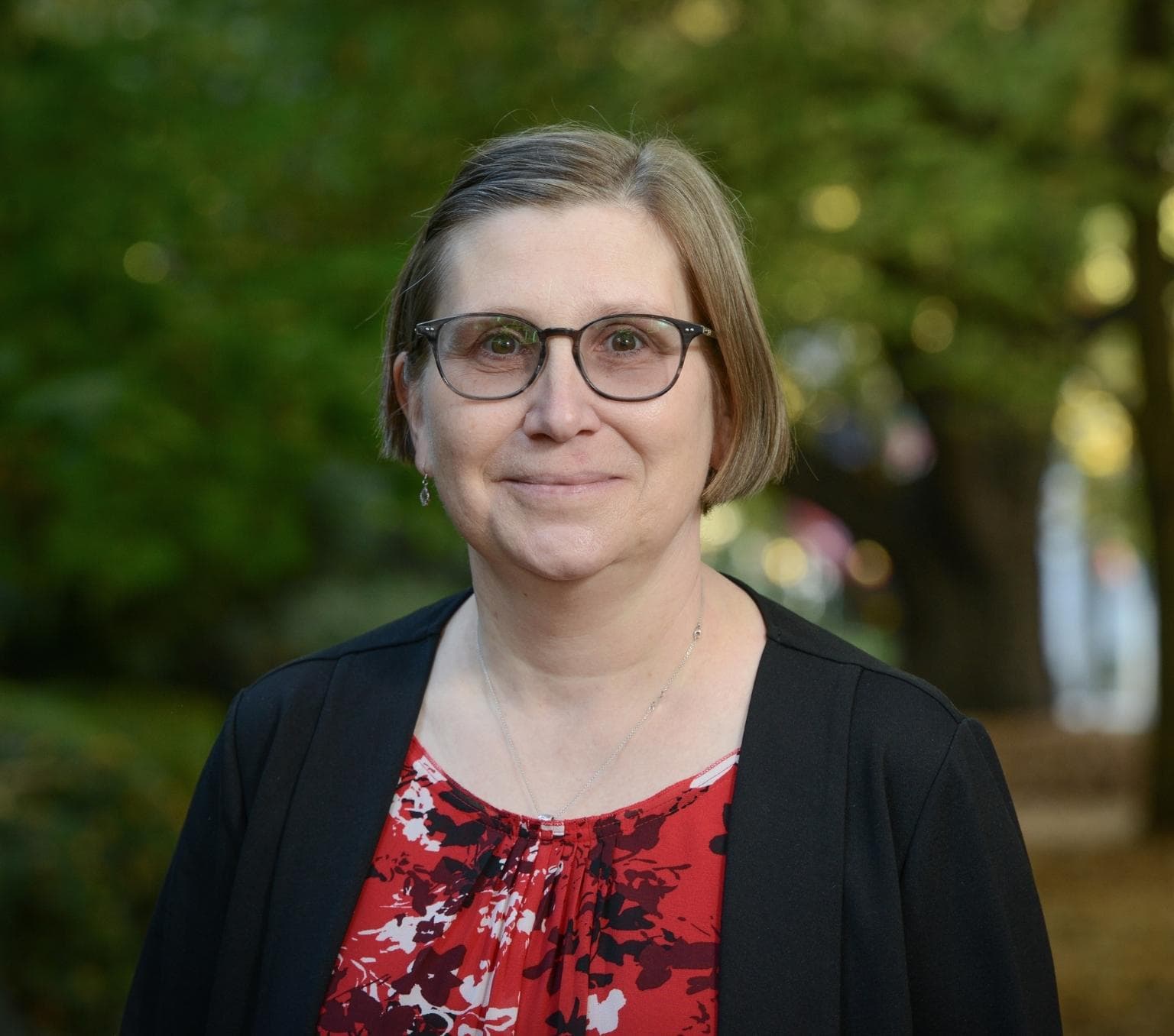 Professor Ute Roessner AM. She is wearing a red, white and black top with a black blazer. There are trees in the background.