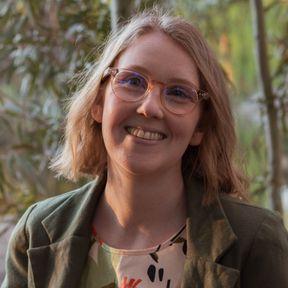 Olivia Congdon smiles at the camera. She has shoulder length blond hair and wears glasses and a khaki-coloured jacket.