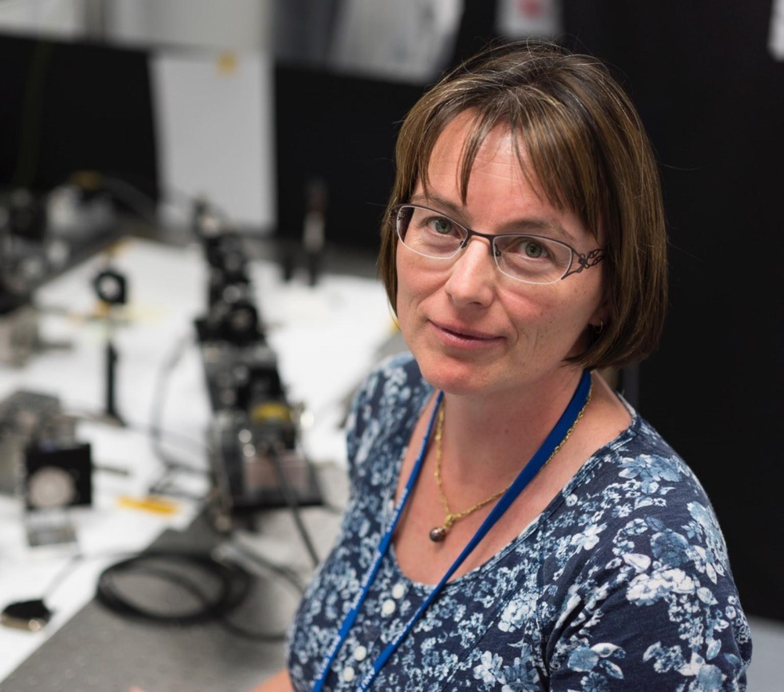 Celine is pictured from the chest up. She is wearring a blue floral top, black rimmed glasses and has a brown bob. Behind he is blurred laboratory equipment