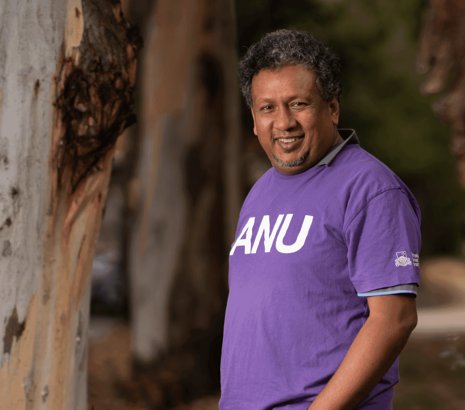 A man is in focus standing next to a tree. He is wearing a purple t-shirt with bold white letters ANU.