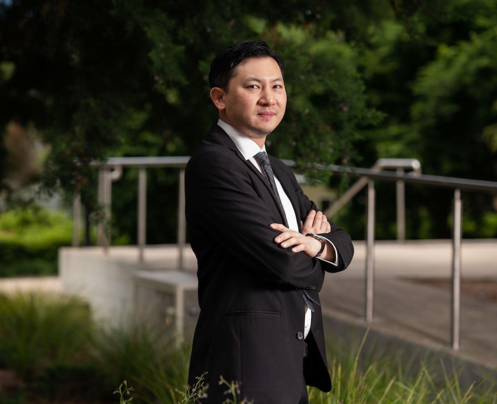 Si Ming Man is dressed in a black suit, with his arms folded. He is standing side on and looking towards the camera.