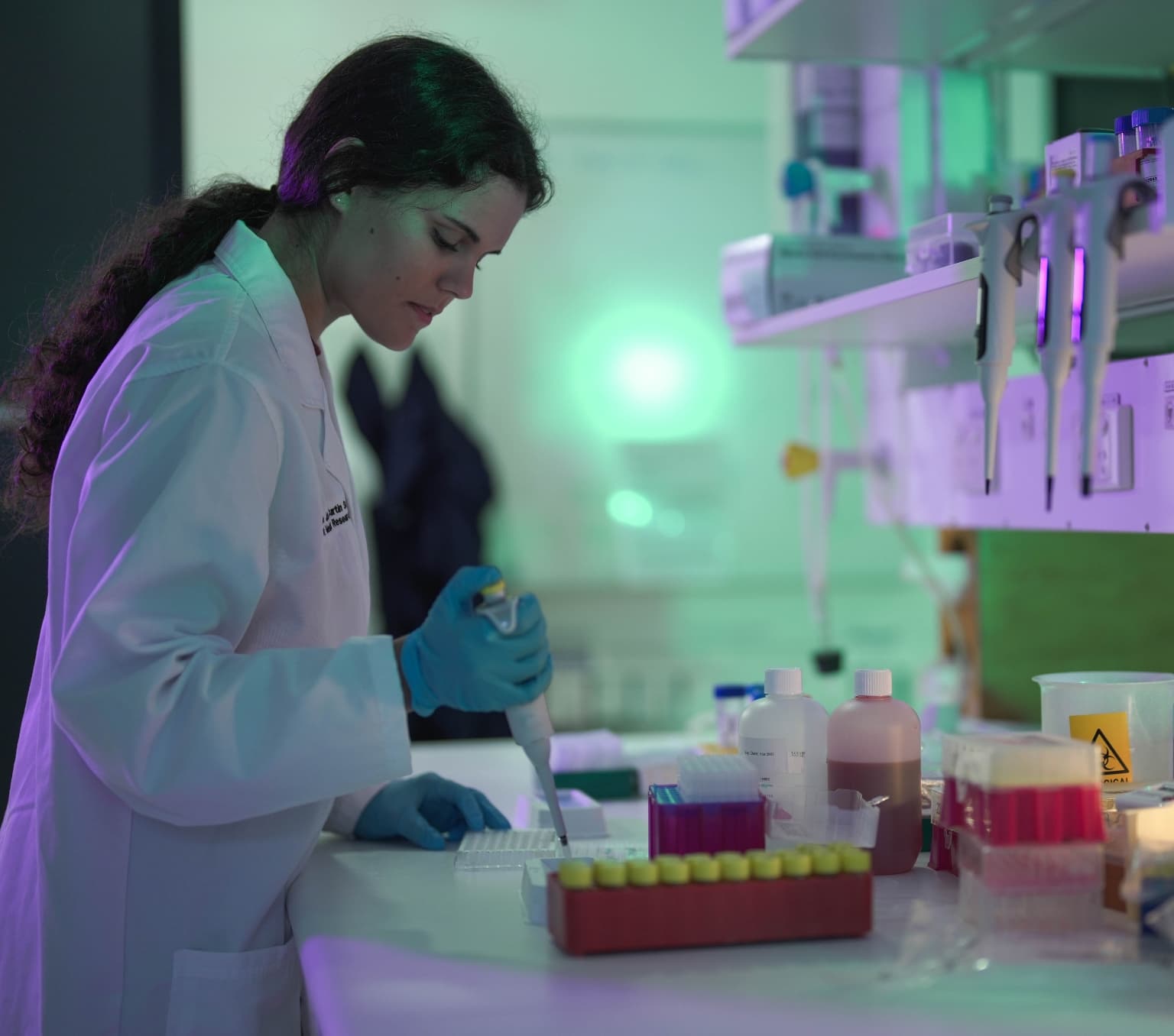 Dr Cynthia Turnbull in the lab. She is wearing a white lab coat and is injecting an unknown substance onto a petri dish.