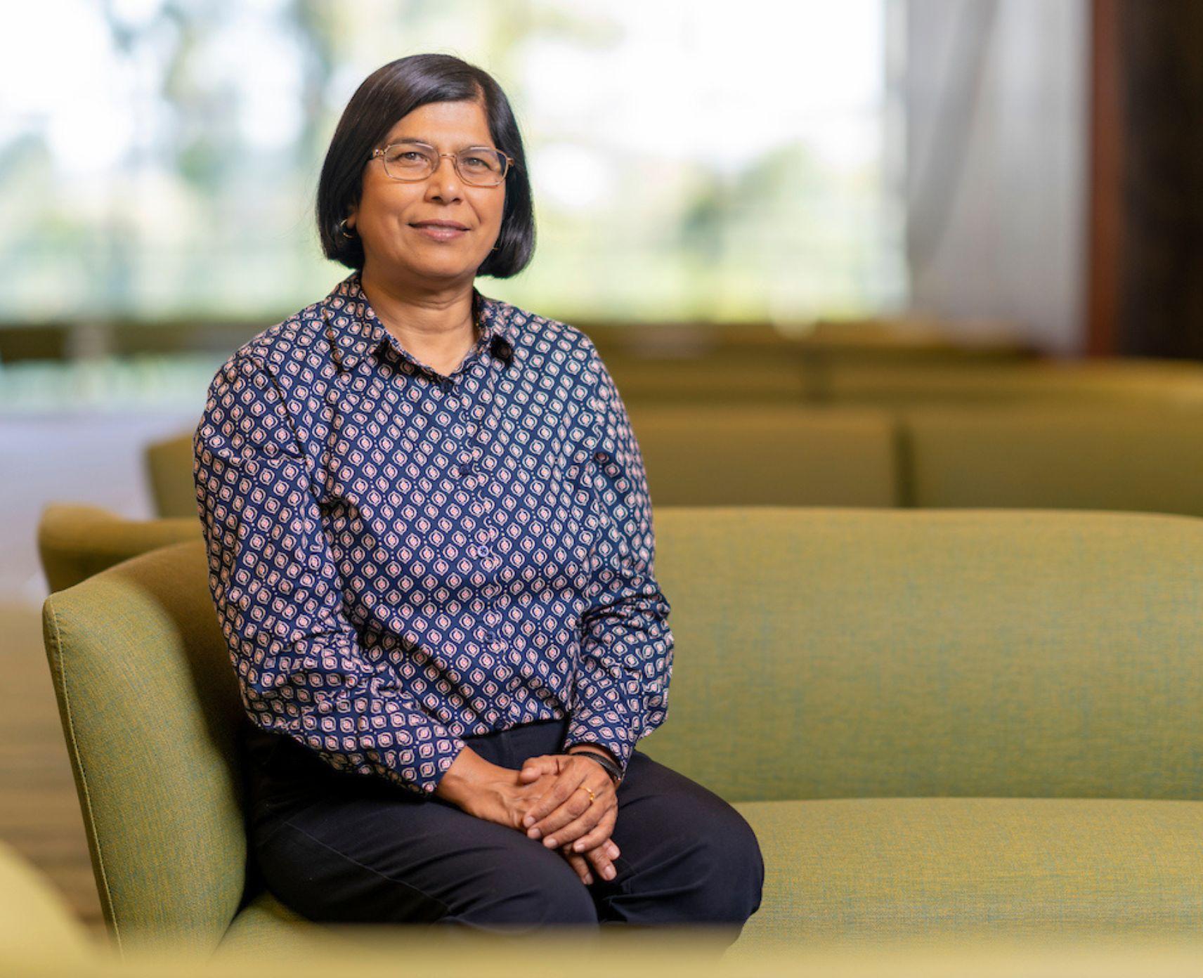 Mahananda sits on a couch similing at the camera. She is wearing a blue shirt.