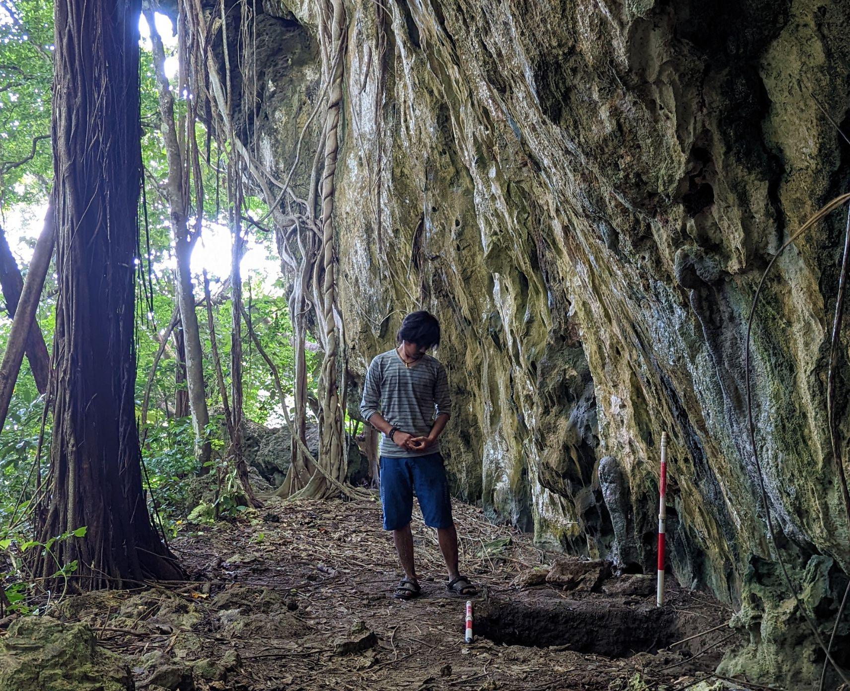 A man stands in front of an archaeological site surrounded by tall trees