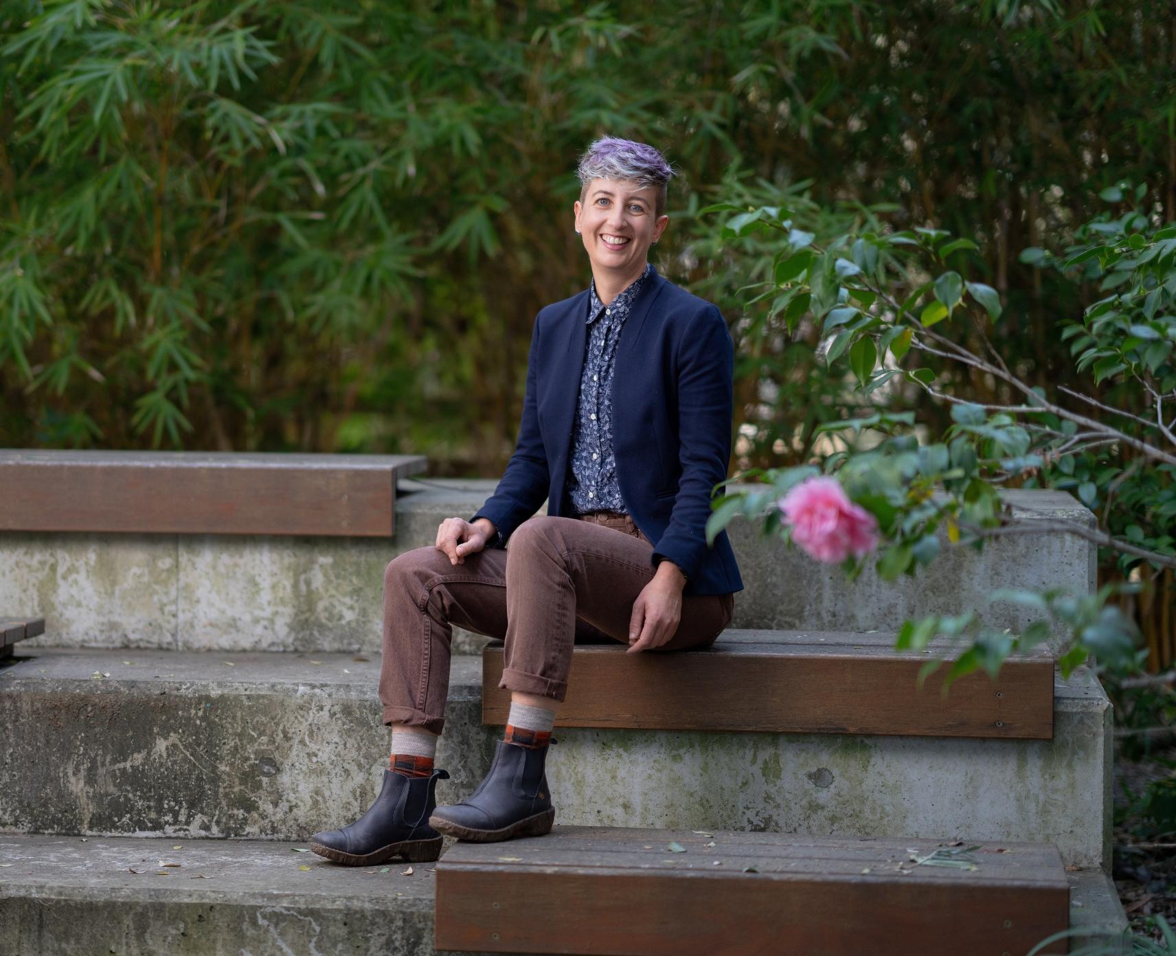 Caroline Schuster, author of a comic book based on her research, smiles at camera seated in front of bamboo. A rose is in the foreground.