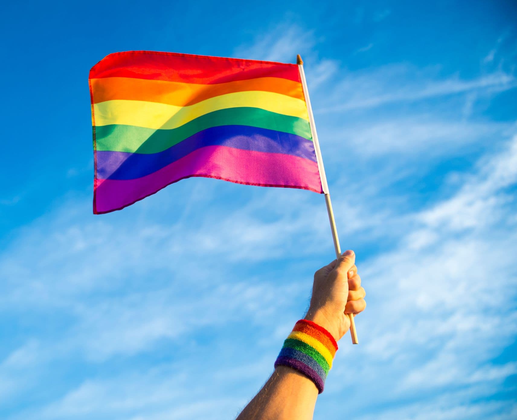 A hand with a rainbow sweatband holds a pride flag up to the sky