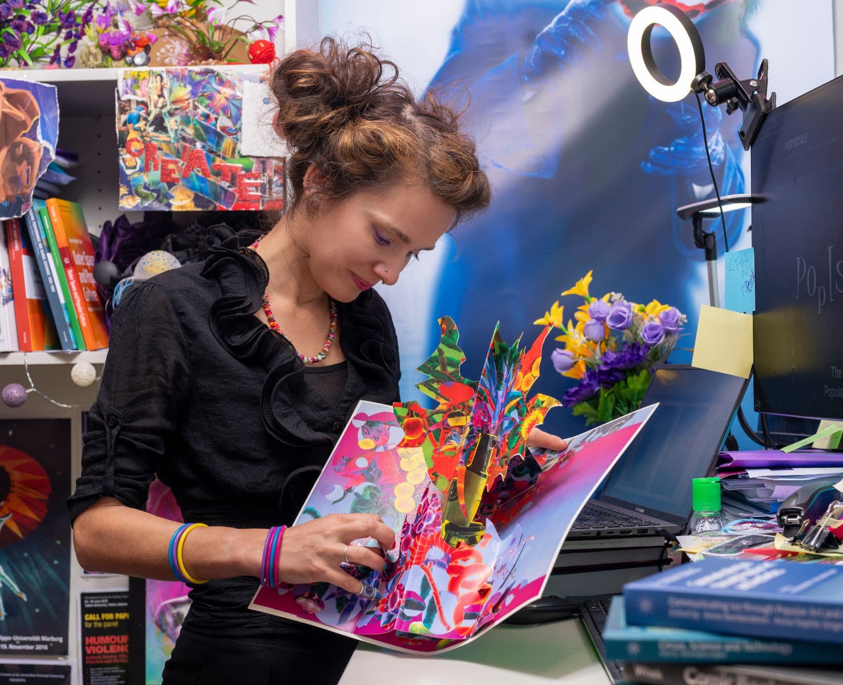 Woman is looking down at an opened book that she is holding, whilst in a colourful room full of posters and books.