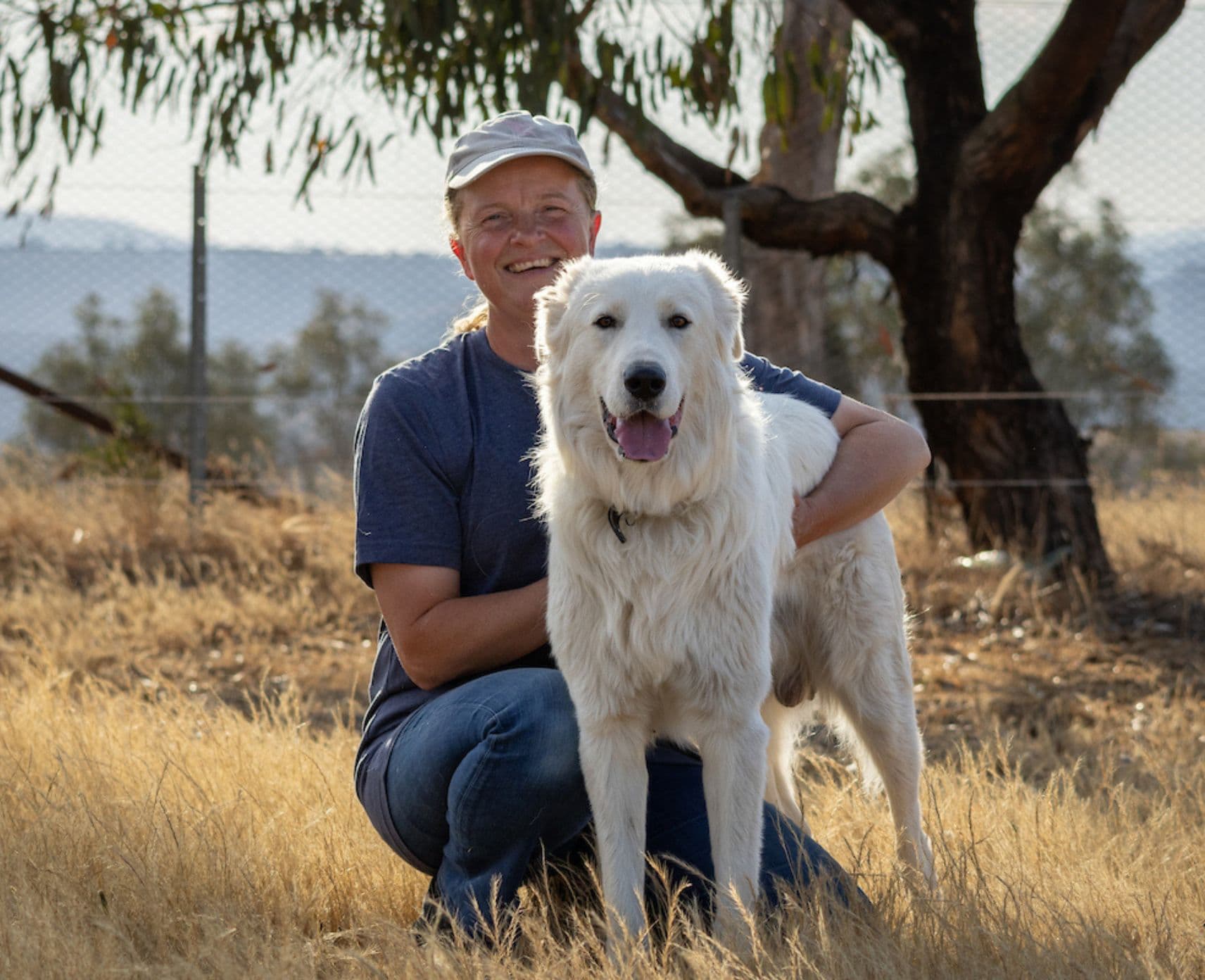 Woman hugging white sheepdog in front of a tree in bushland.