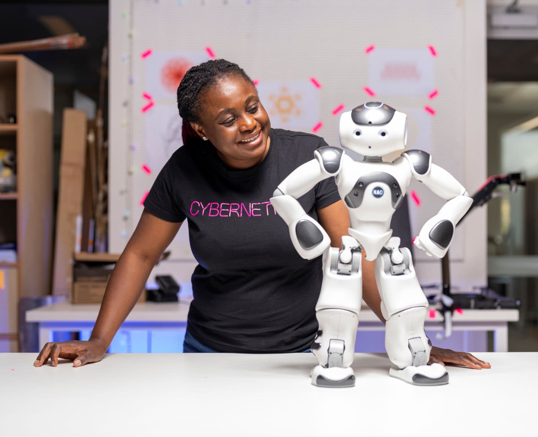 A woman looks at Nao, a small, white robot that is posing with its hands on its hips.