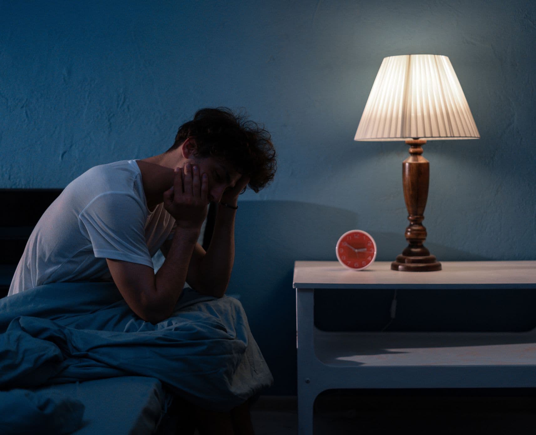 Man sitting on the edge of the bed looking exhausted. he's got his head in his hand.