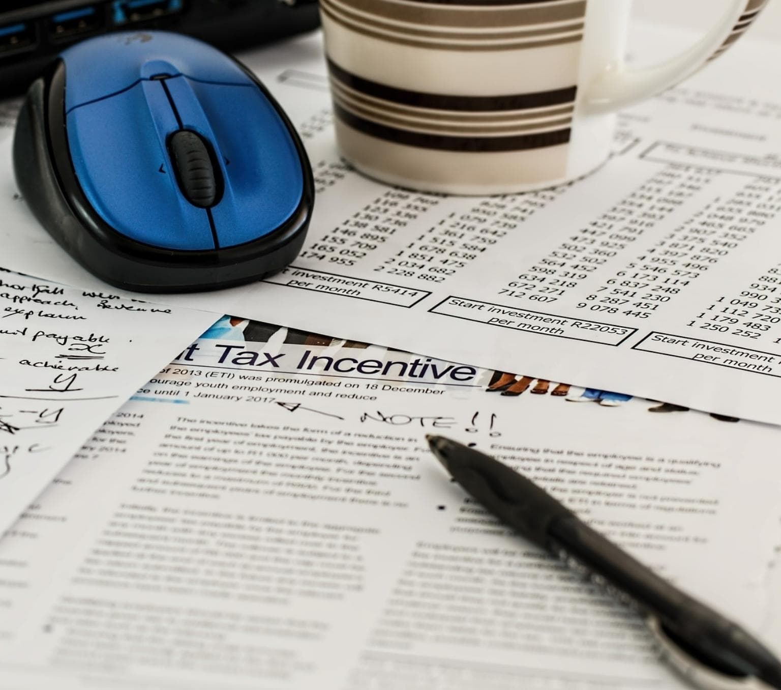 A pen, a computer mouse and coffee cup sit on a papers that have numbers written on them.