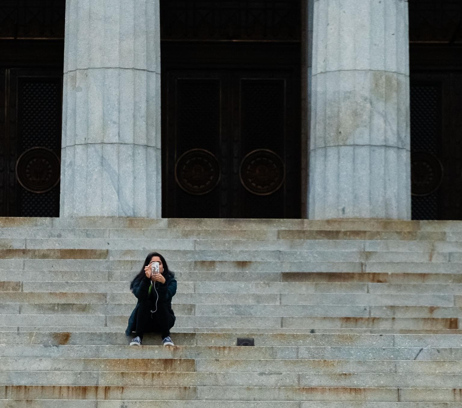 young person looking at their phone and sitting on a set of sandstone steps with large round columns behind. There is no one else in the image
