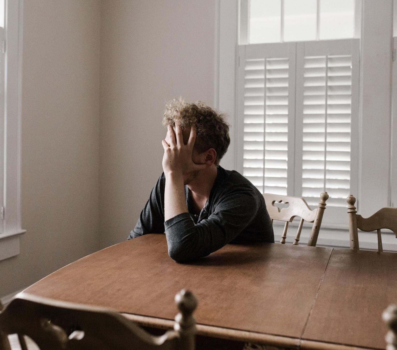 profile of a man sitting at a woodem table. His hand is held to his face and obscures his face. the room he is in is white.