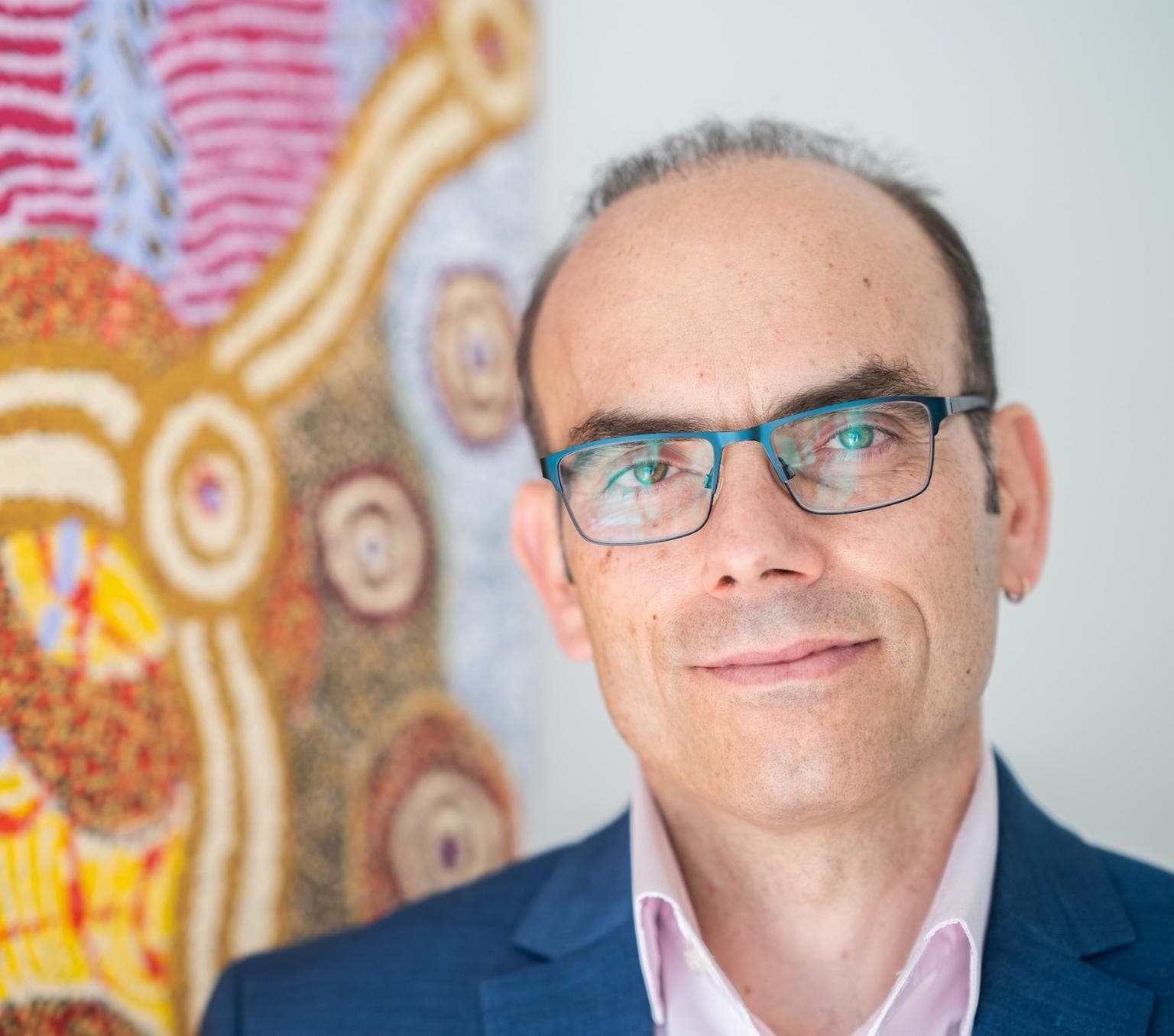 A man is wearing glasses and pictured from the neck up. He is standing next to a brightly coloured pink and yellow artwork