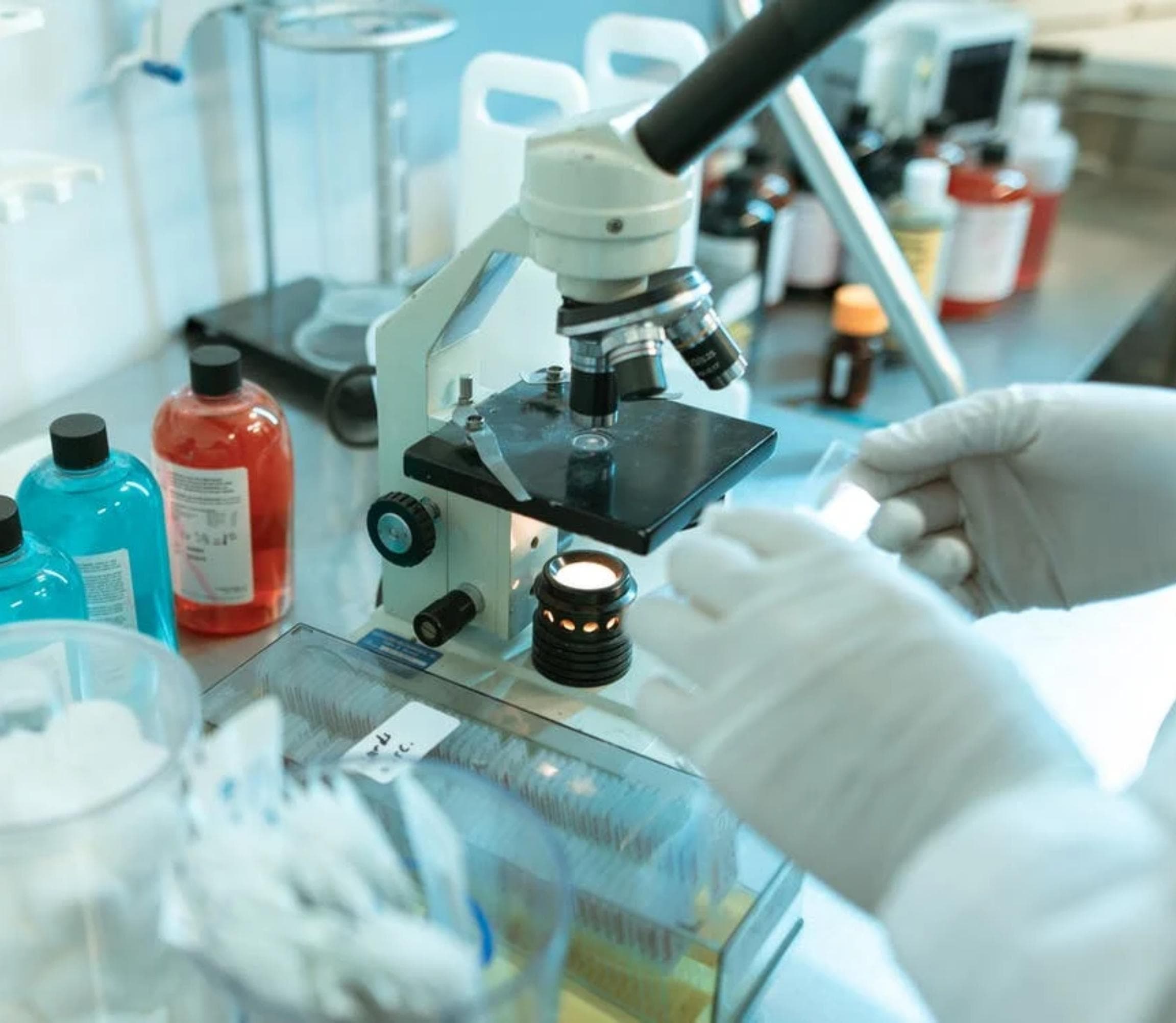 close up of a microscope surrounding by various laboratory equipment including glass slides and bottles of chemicals. The gloved hands of a person are holding a glass slide.