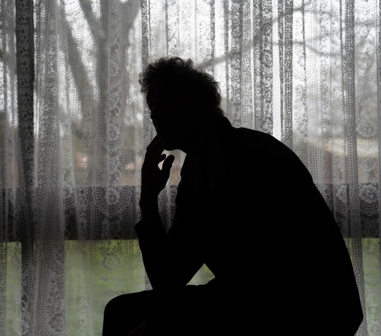 A shadowy figure sits by a window with the curtains closed. The person can't be identified.