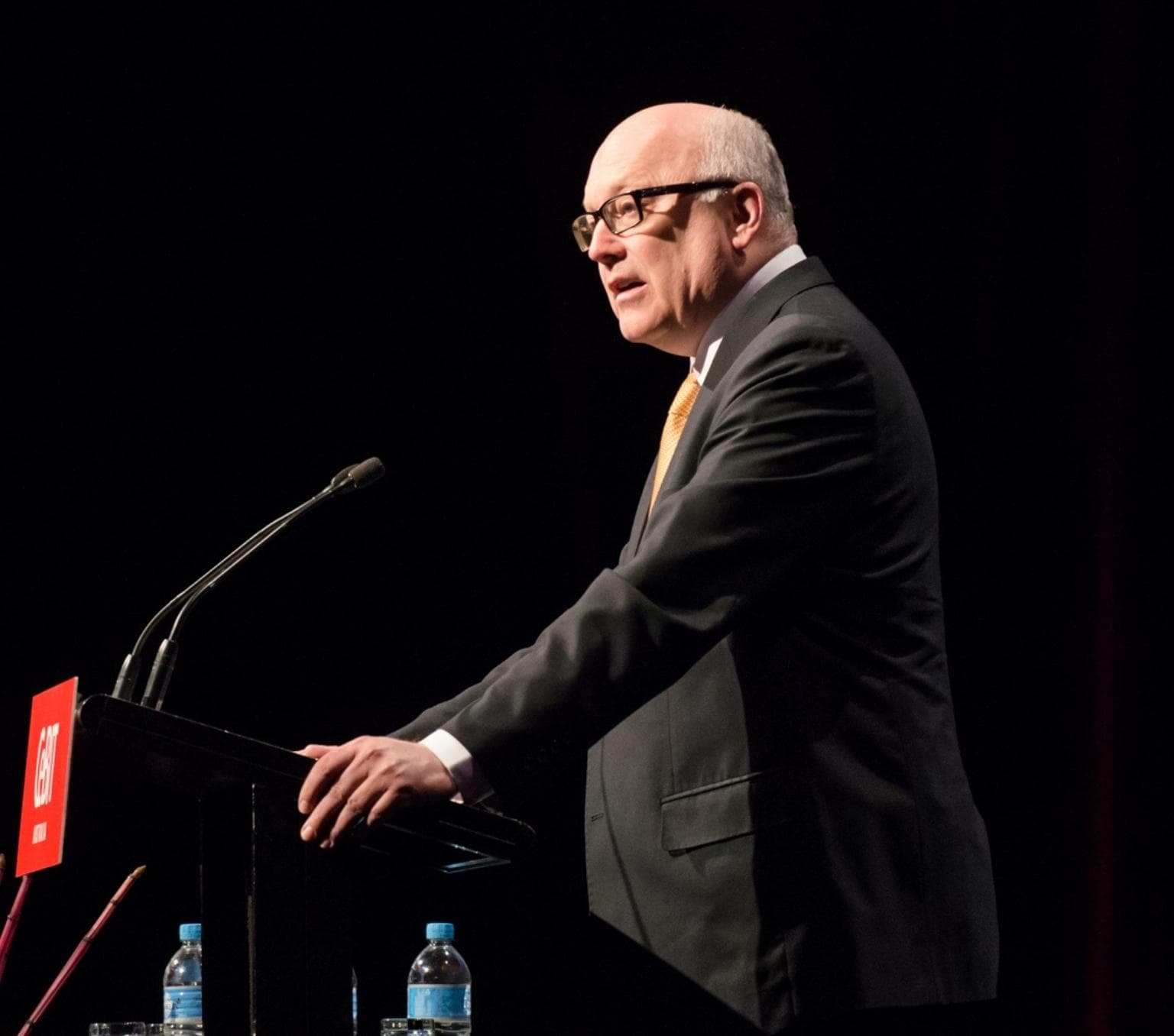 George Brandis is standing side on to the camera. He is wearing a black suit and black rimmed glasses. He is holding onto the side of a lecturn andis mid speech