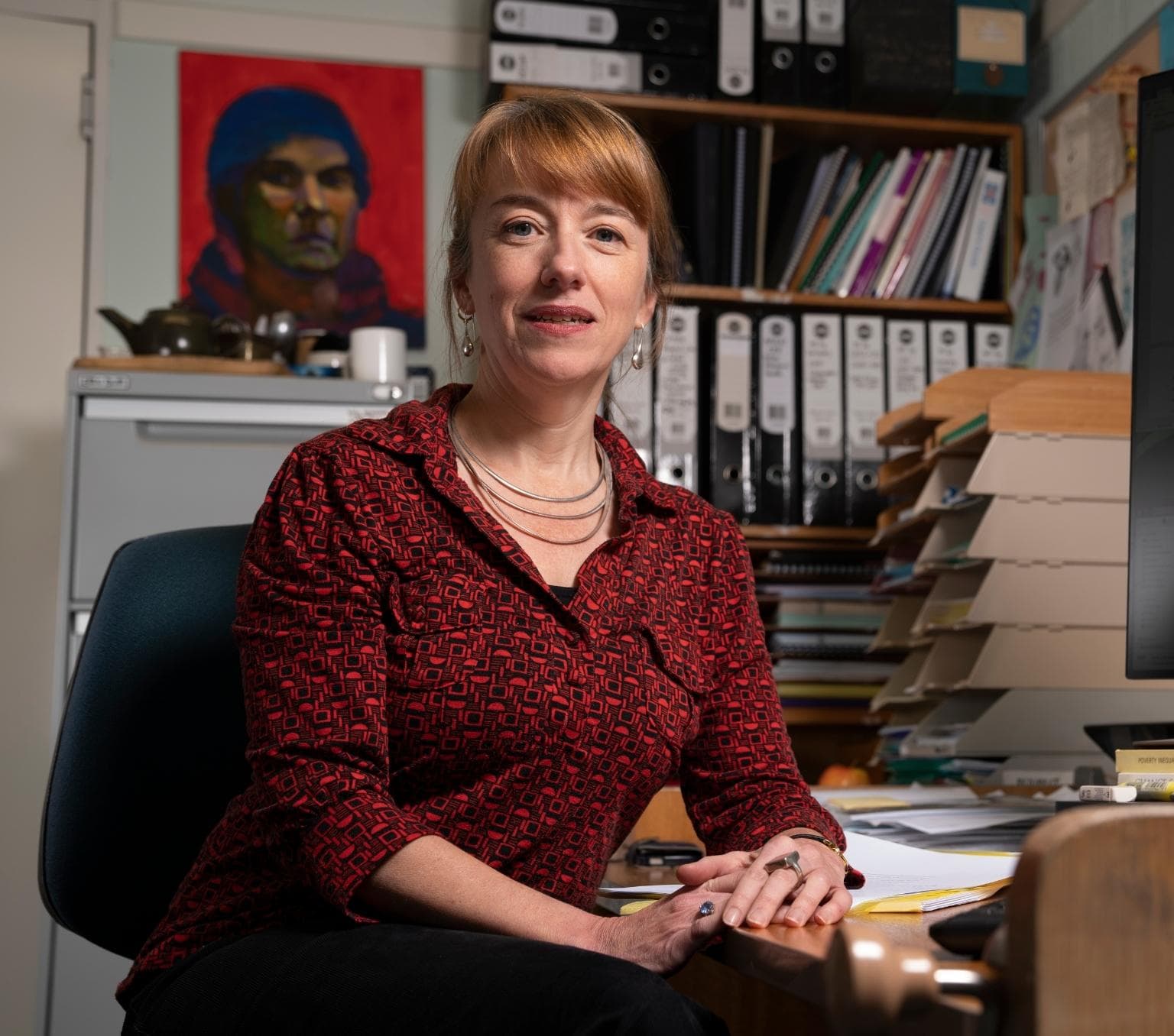 Emily Banks, wearing a red patterned shift, sits at a desk.