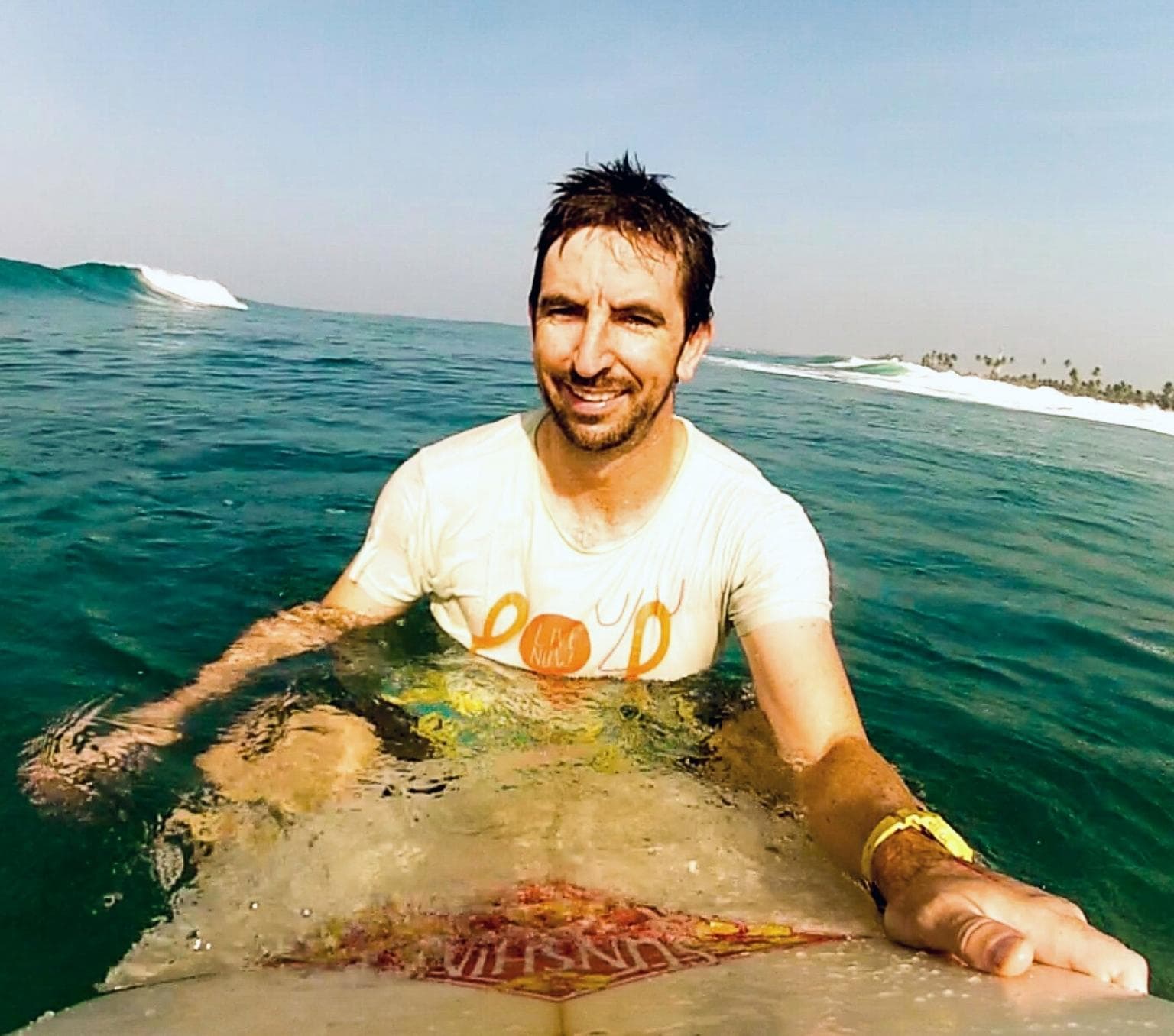 Nicholas Taylore is smiling at the camera. He is wearing a white T-shirt and sitting on a surfboard in the ocean. There are small waves behind him.