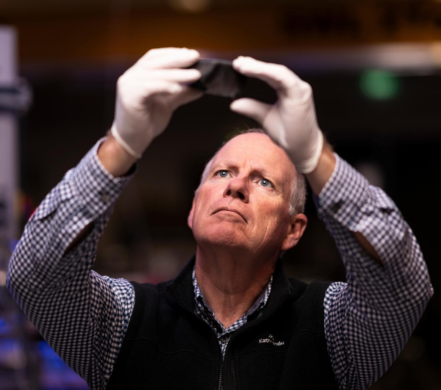 Trevor Ireland wears a blue and white collared shirt and a black vest. He is holding a black rock slightly above his head in front of him and looking at it