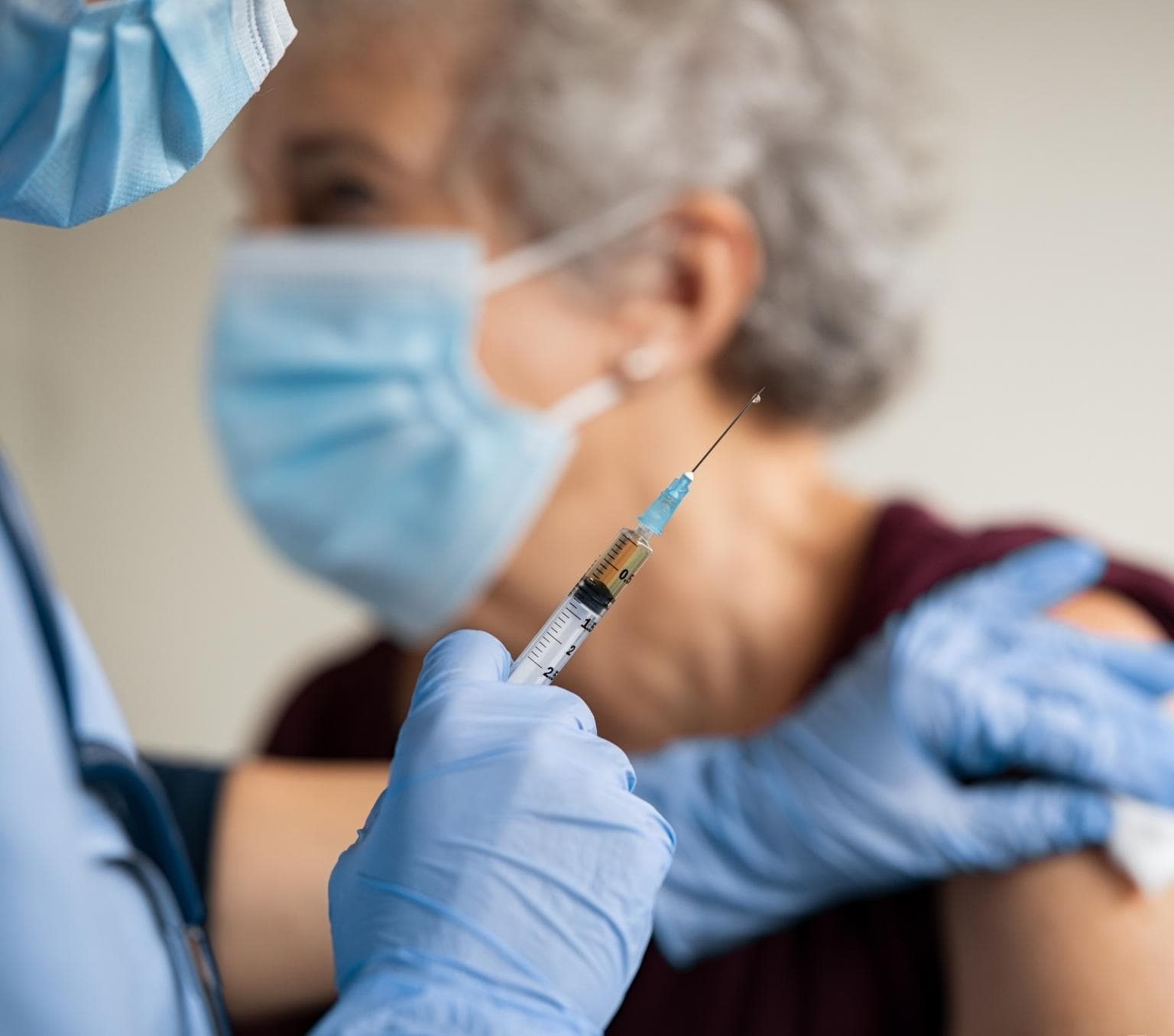 A person wearing blue gloves holds a needle in their right hand. Their left hand touches the bare arm of a woman, who is blurred in the background.