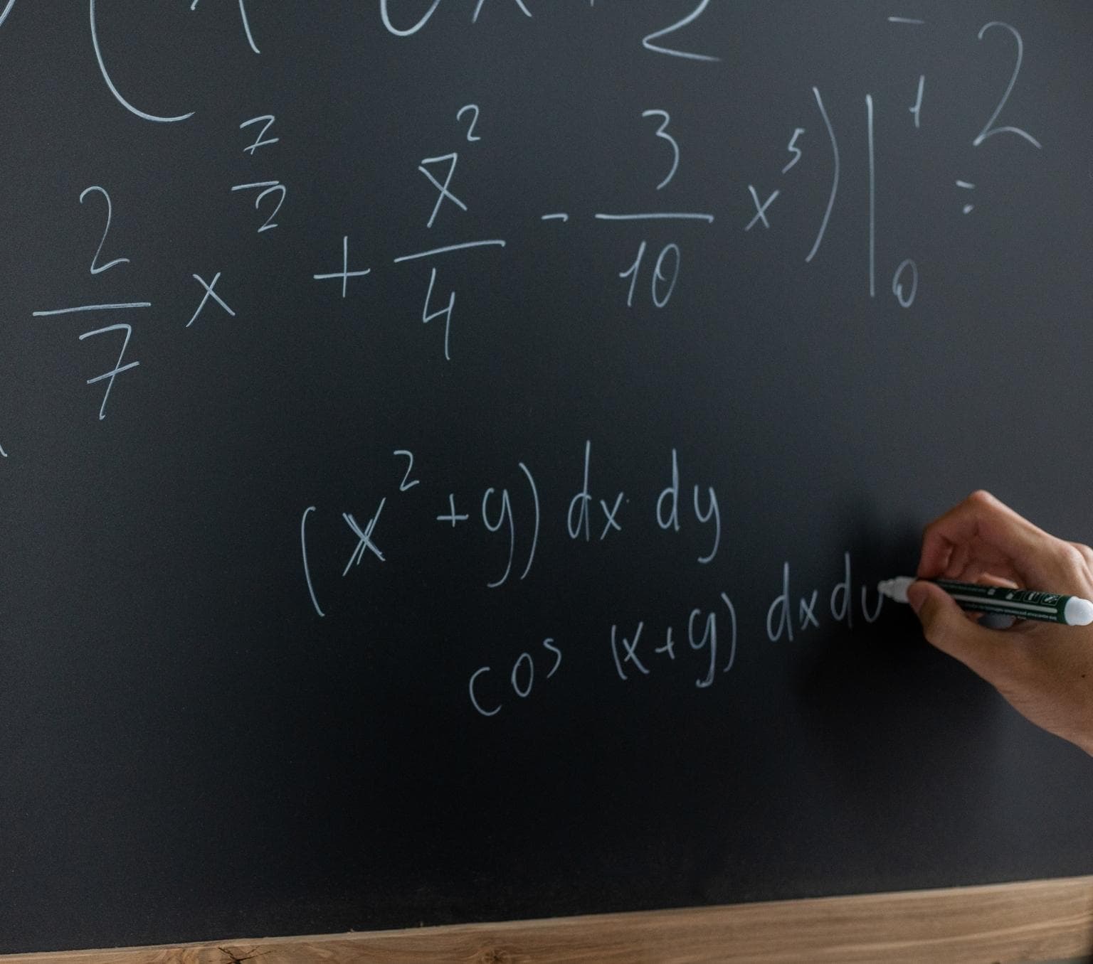 black board with maths equations written in white marker. a hand is picted at the bottom of the frame writing a maths equation.
