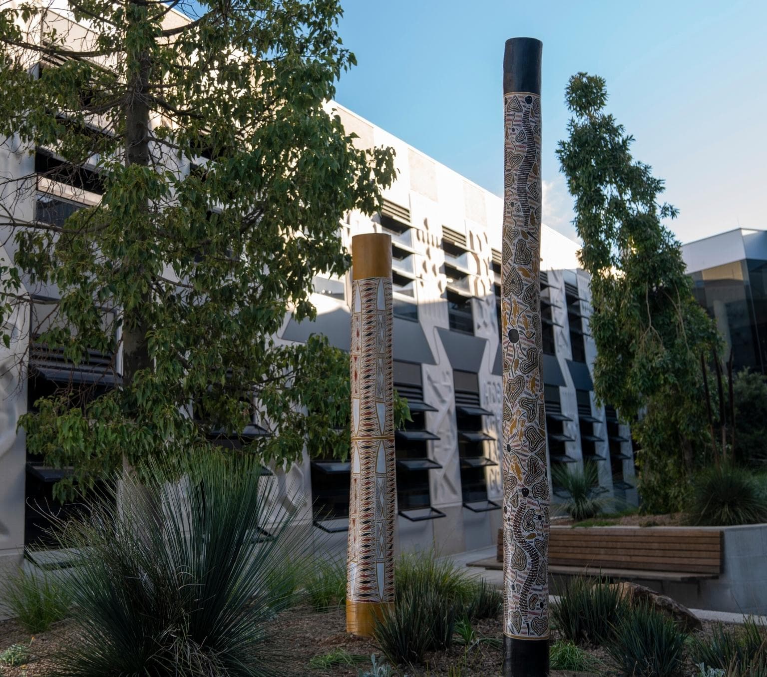 Memorial poles made of wood and painted in black, yellow and qhite indigenous artwork are located in a small garden ext to a tree. The garden is in front of a white building. There is blue sky in the background.