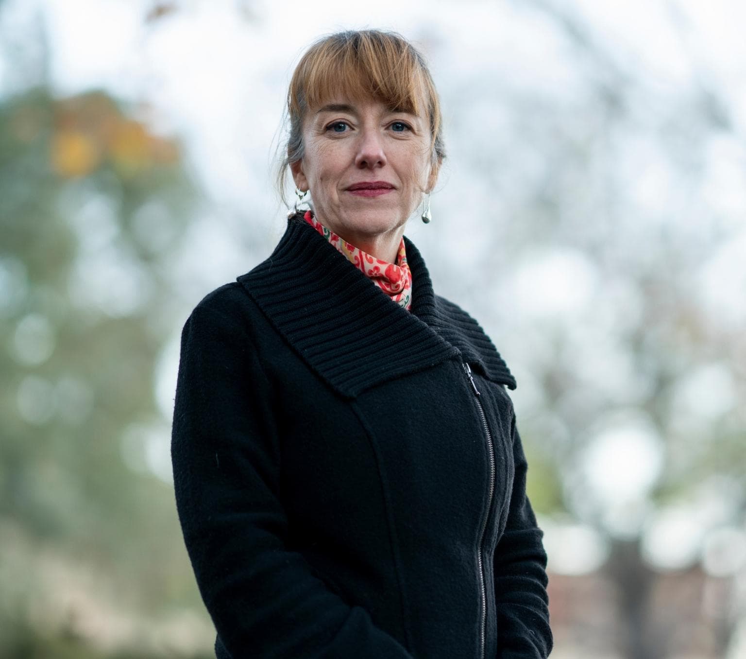 Emily Banks is standing outside in front of a blurred background of trees and sky. She is wearing a black coat with a red patterned scarf and looking at the camera