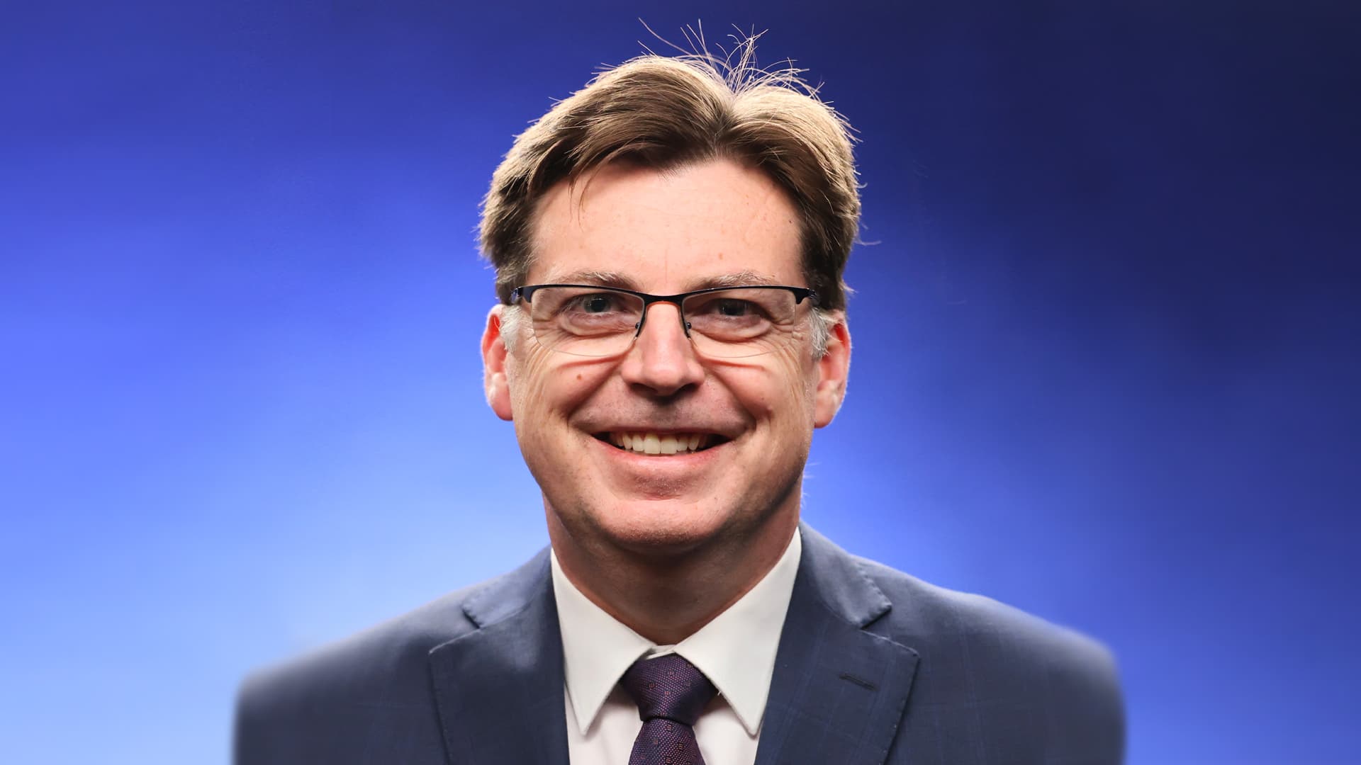 Dave is pictured from the chest up wearing a blue suit and purple tie. He is in front of a blue background and smiling at the camera