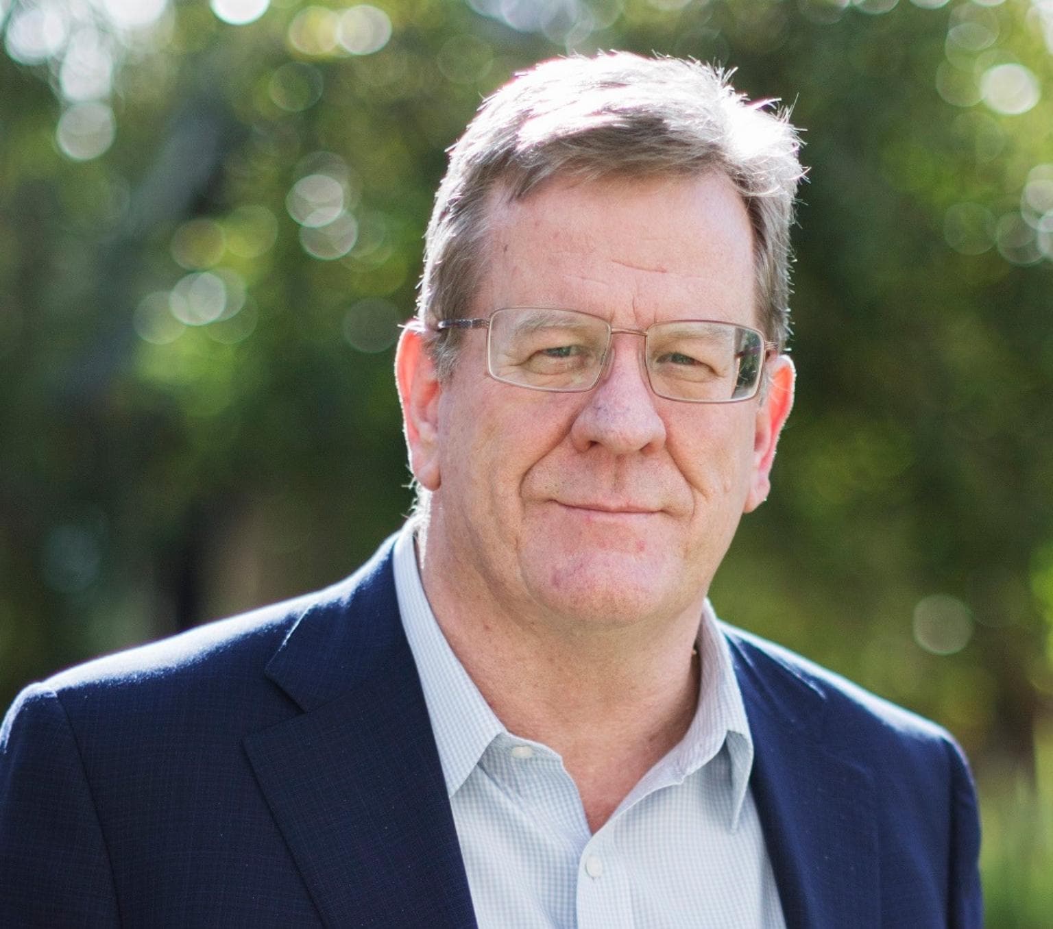 Brendan is pictured from the chest up with a neutral expression. He is wearing glasses and a blue suit