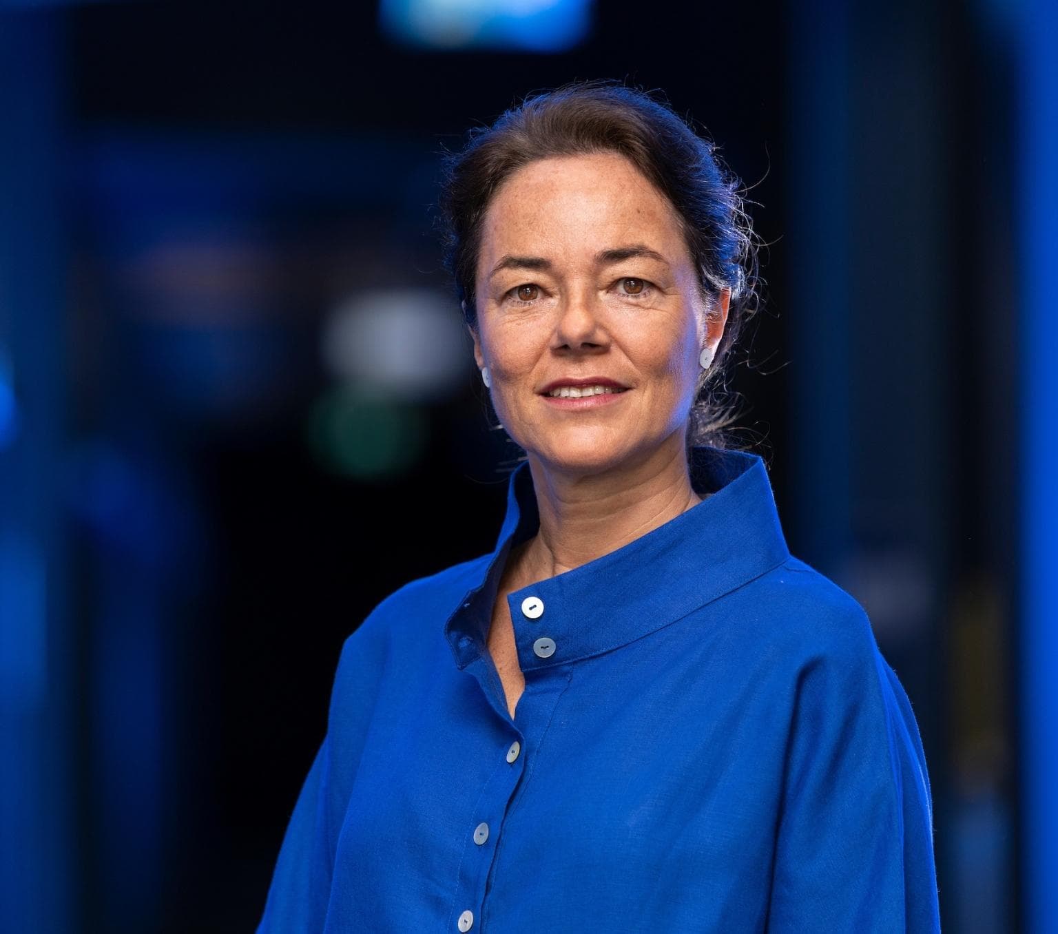 Carola is wearing a blue shirt and smiling at the camera. She has brown hair that is tied up