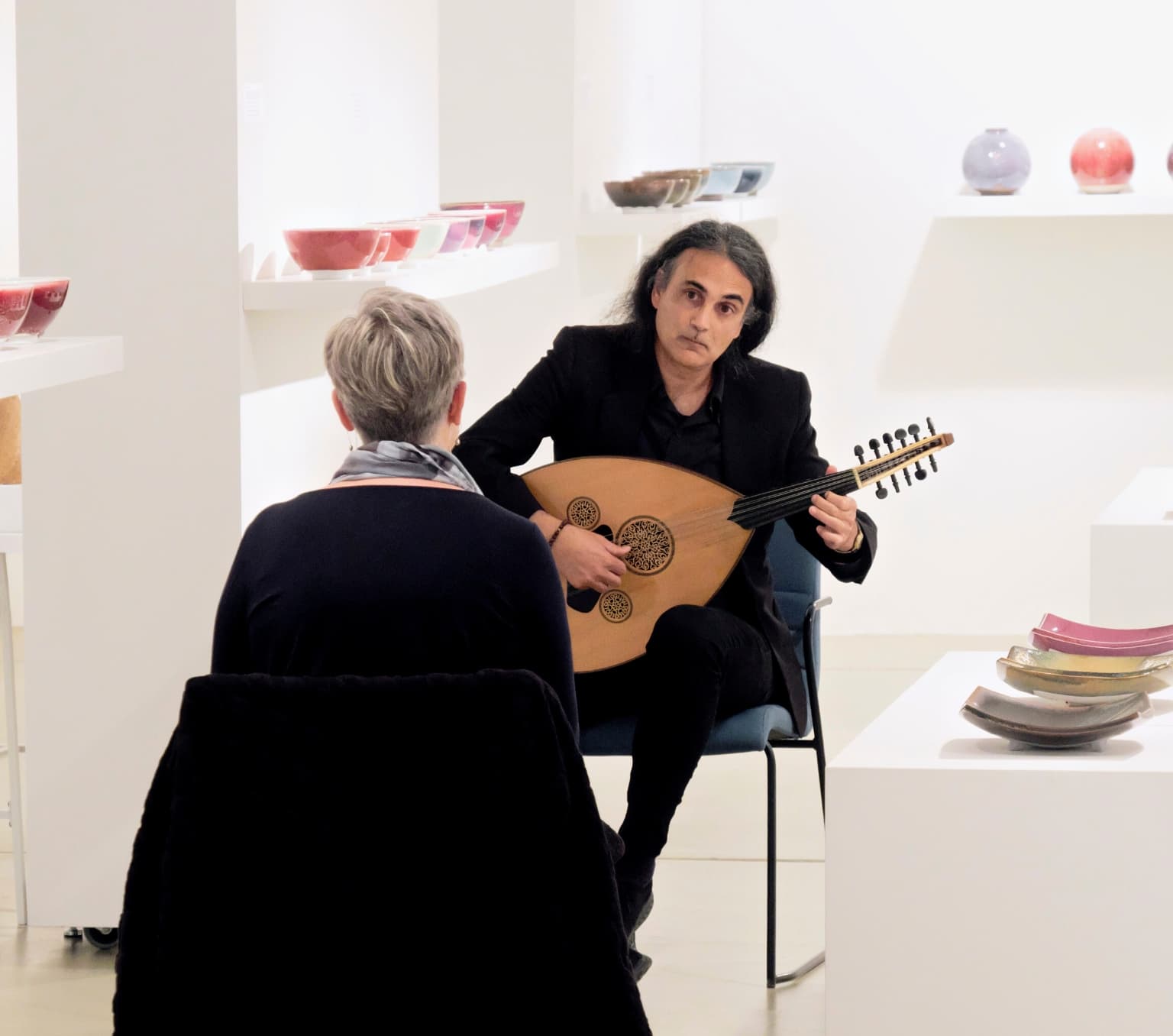 a man is sitting in a chair and playing a round wooden instrment similar to a guitar for a woman whose back is facing the camera