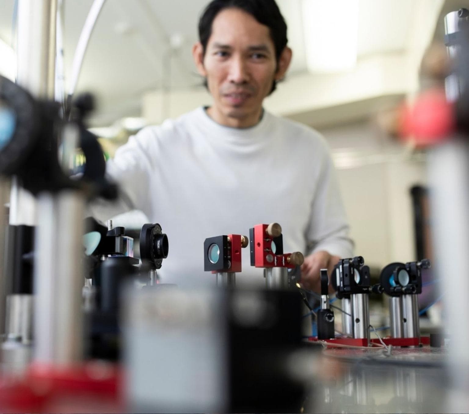Syed is wearing a white shirt. He is standing behind a range of technical objects in a laboratory