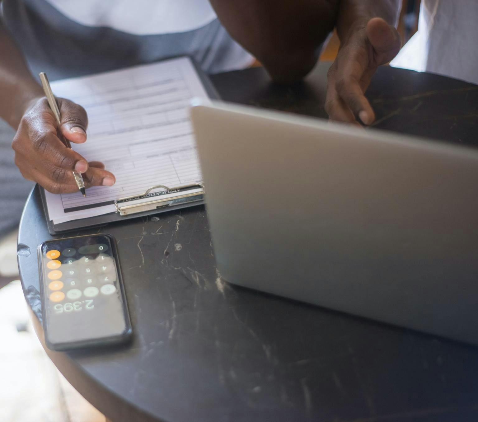 Close up of a desk with a person's hands holding a pen. There is also a laptop, phone and clipboard on the desk