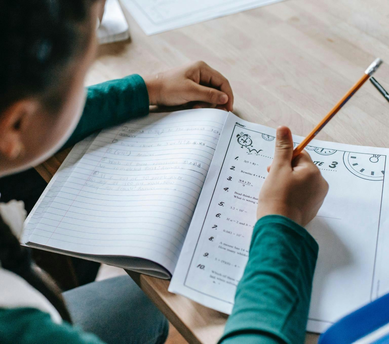 image is taen over the shoulder of a child wearing a green long sleeve shirt. The child is holding a yellow pencil and writing in a maths worksheet