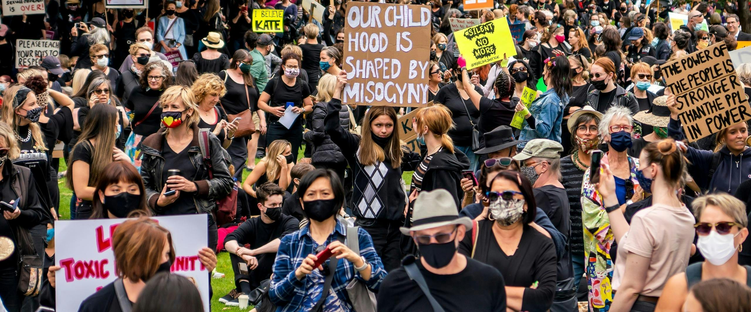 A large group of people stand outside. In the centre a woman holds up a cardboard sign the message 'our childhood is shaped by misogyny' in large white letters.