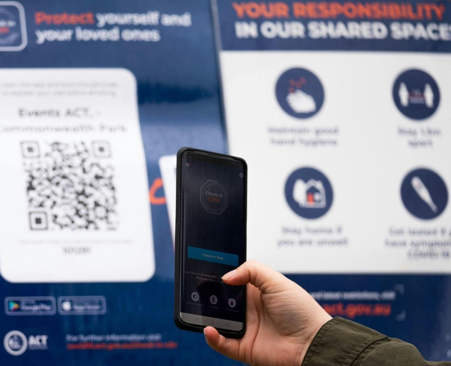 close up photo of a woman's hand holding a phone infront of Canberra COVID-19 checkin posters. The phone displays the Canberra check-in app.