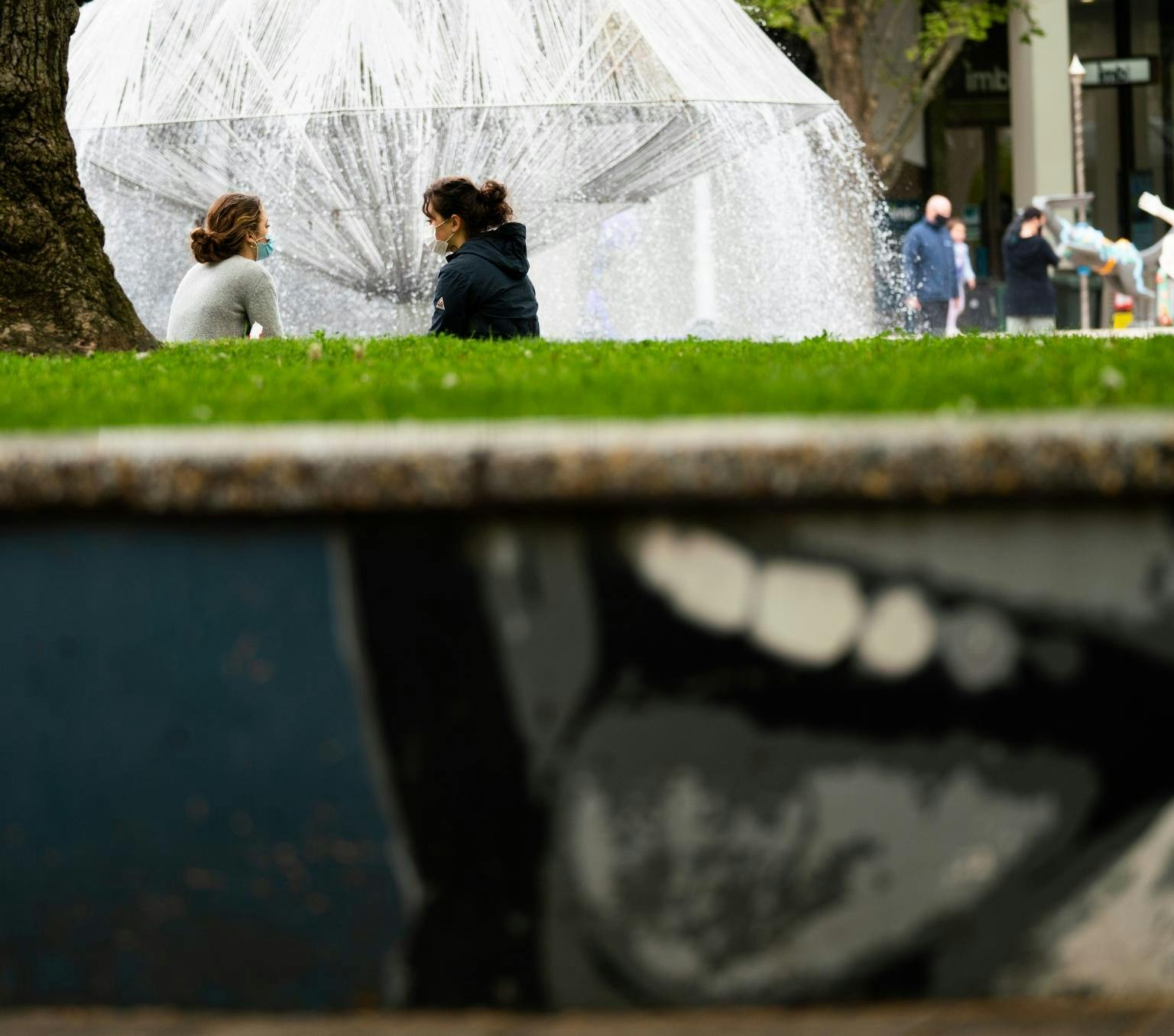 Two people wearing masks sit on a bench and talk to each other. There is a fountain in the background and a painting of a mouth on a wall in the foreground.