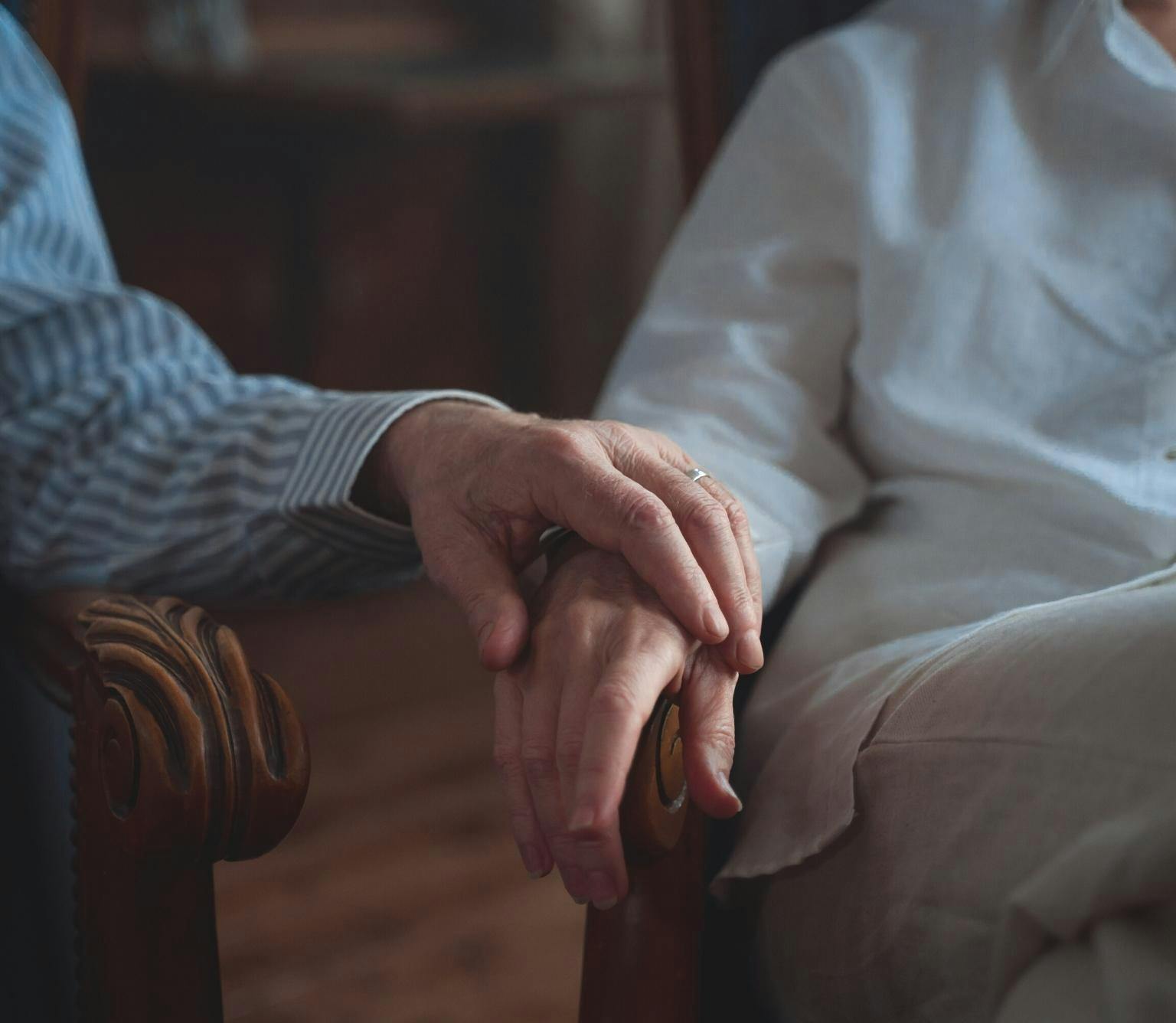 close-up of a hand place on top of another persons hald which is resting on an arm chair. Both hands are slightly wrinkled.