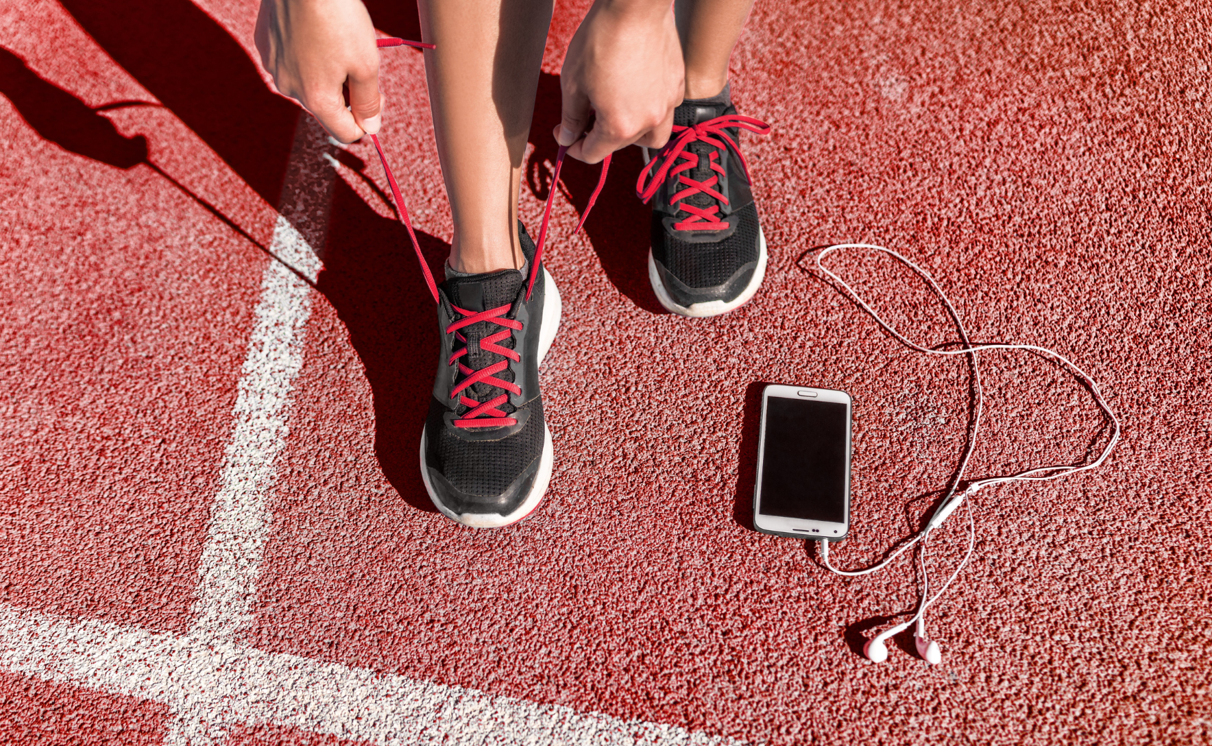 A runner on an athletic track pulls on the laces of their untied running shoes. Next to their feet is a smartphone and headphones.