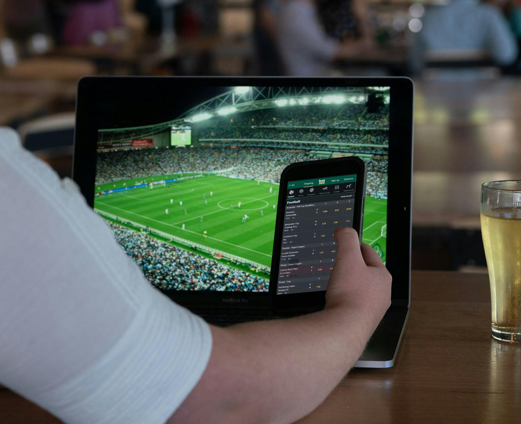 Young woman holds her phone up whilew watching sport on her laptop. She's wearing a white top.