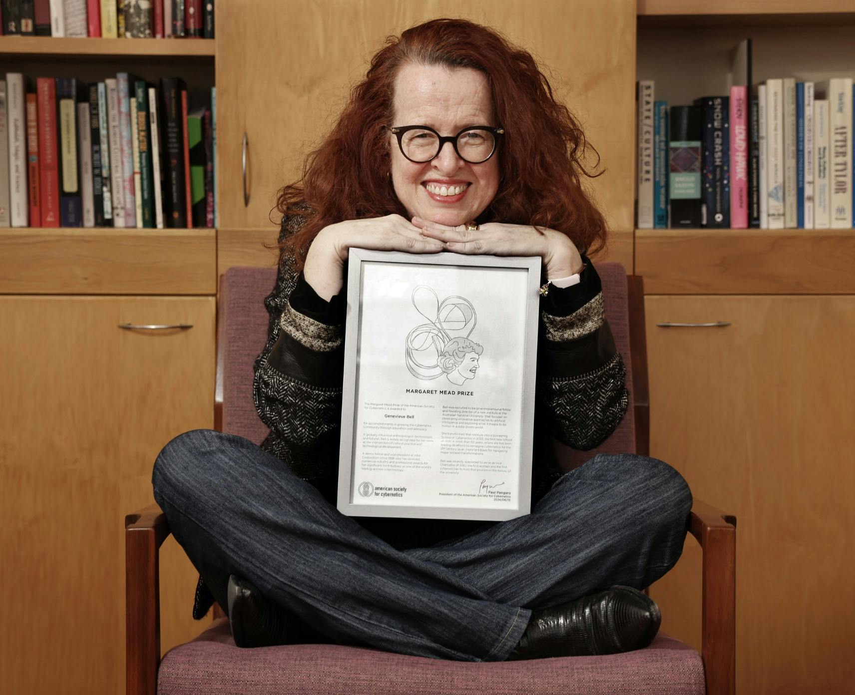 Genevieve Bell sits cross-legged in a chair and holds her Margaret Mead award on her lap.